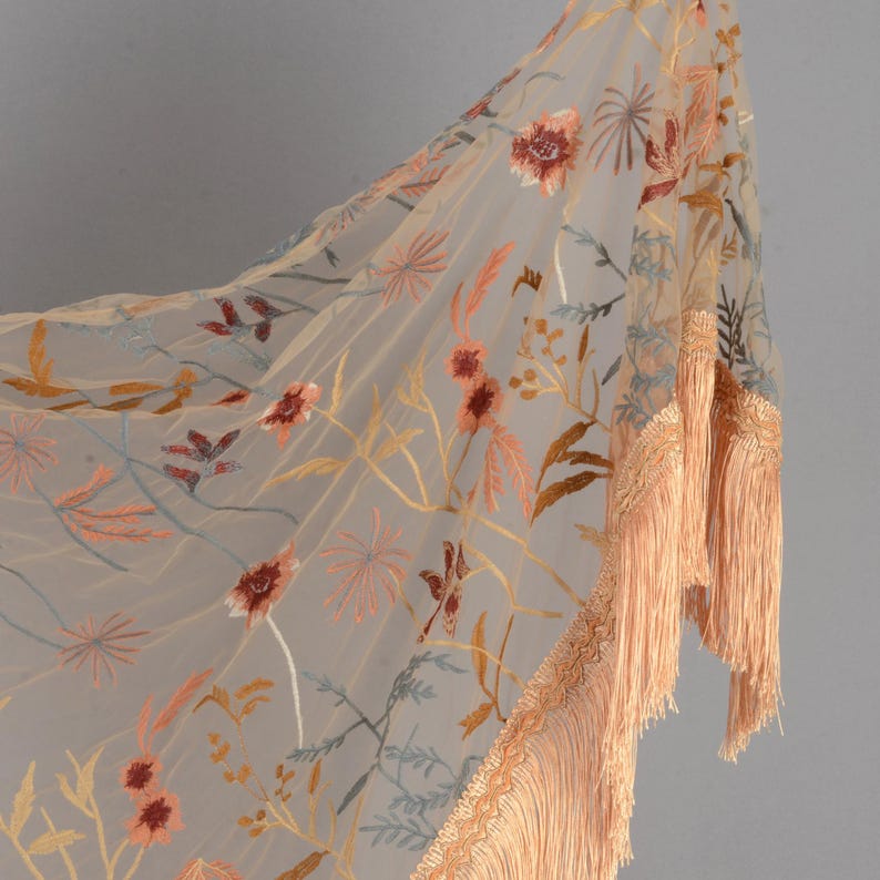 May include: A sheer, beige shawl with embroidered floral patterns in shades of red, orange, and blue. The shawl features a fringe trim in a matching peach color, adding a decorative element to the garment.