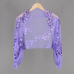 Op de afbeelding: Een lavendelkleurige, met pailletten bezette shrug met lange mouwen en een gerimpelde zoom. De shrug is gemaakt van een transparante stof en is versierd met kleine, iriserende pailletten.