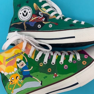 Pode incluir: Um par de sapatos de lona verde e branco de cano alto com desenhos pintados à mão. Um sapato apresenta um personagem de desenho animado com uma espada e um cão. O outro sapato apresenta um personagem de desenho animado com uma guitarra e um pássaro. Os sapatos têm o texto "Victoria Since 1915" no lado.