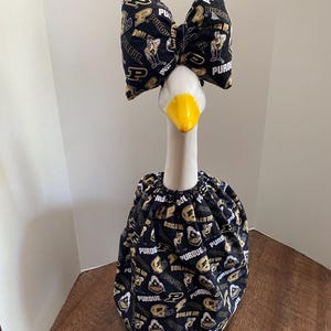 May include: A white goose statue wearing a black fabric outfit with a repeating Purdue University logo, text reading Boiler Up, and a matching large fabric bow attached to the head. The goose has a bright yellow beak and a gathered neck collar.