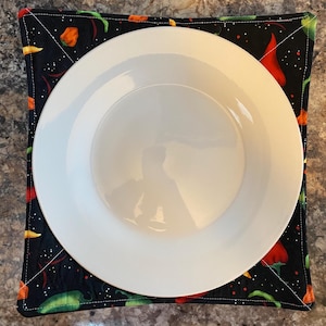 May include: A white ceramic plate sits on a black fabric hot pad with a chili pepper pattern. The hot pad has a white border and is quilted.