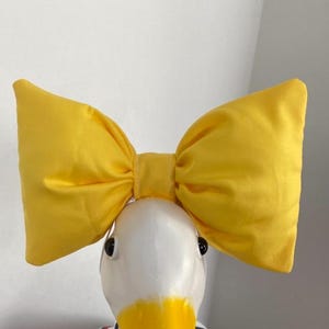 May include: A bright yellow fabric bow attached to a headband. The bow is large, puffy, and features a central knot detail. It is positioned on top of a white, smooth, duck-shaped display head that has black eyes and a yellow beak.