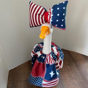 May include: A white goose figurine wearing a fabric outfit featuring a red, white, and blue patriotic patchwork pattern with stars and stripes. The goose has a bright orange beak and wears a matching oversized bow on its head with the same print.