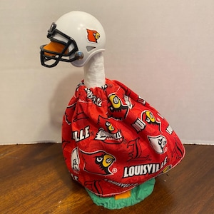 May include: A decorative goose figurine wearing a white football helmet with a black face mask and a red fabric outfit featuring the Louisville Cardinals logo and text. The goose has a white neck and stands on a green base with an orange beak.
