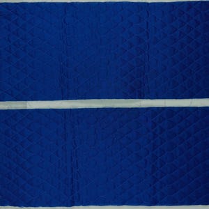 K&ouml;nnte beinhalten: Zwei rechteckige St&uuml;cke aus blauem, gestepptem Stoff mit einem Rautenmuster. Der Stoff hat ein strukturiertes Aussehen und rohe Kanten. Die Teile sind horizontal angeordnet, durch einen wei&szlig;en Raum getrennt, vor einem wei&szlig;en Hintergrund.