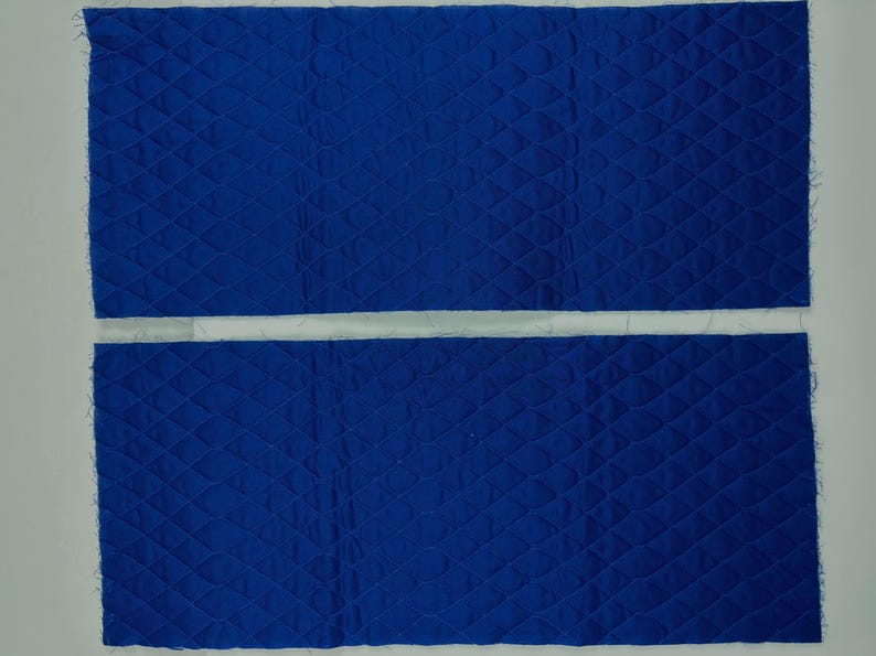 K&ouml;nnte beinhalten: Zwei rechteckige St&uuml;cke aus blauem, gestepptem Stoff mit einem Rautenmuster. Der Stoff hat ein strukturiertes Aussehen und rohe Kanten. Die Teile sind horizontal angeordnet, durch einen wei&szlig;en Raum getrennt, vor einem wei&szlig;en Hintergrund.