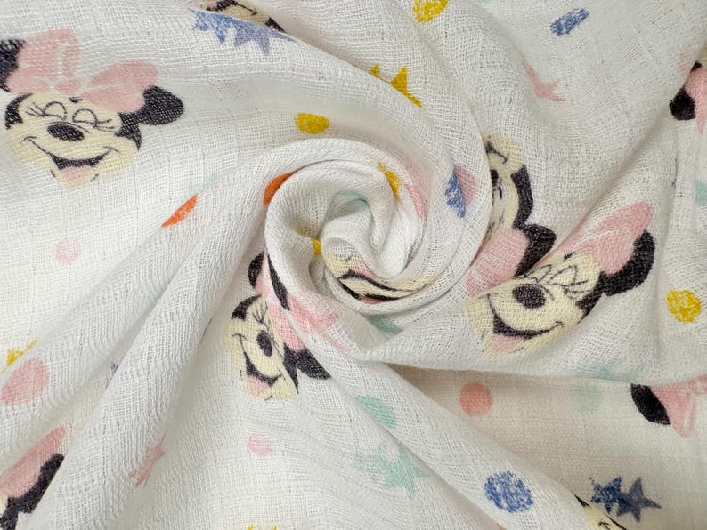 Op de afbeelding: Witte stof met een herhaald patroon van Minnie Mouse-gezichten, roze strikken en kleurrijke sterren en stippen. De stof is zacht en getextureerd, met een licht gekreukte uitstraling. Het ontwerp is speels en geschikt voor kinderartikelen.