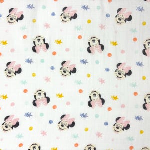 Op de afbeelding: Witte stof met een herhaald patroon van Minnie Mouse-gezichten, roze strikken en kleurrijke stippen en sterren. De Minnie Mouse-gezichten zijn zwart en wit met roze strikken. De stippen zijn oranje, roze en blauw, en de sterren zijn geel en blauw.