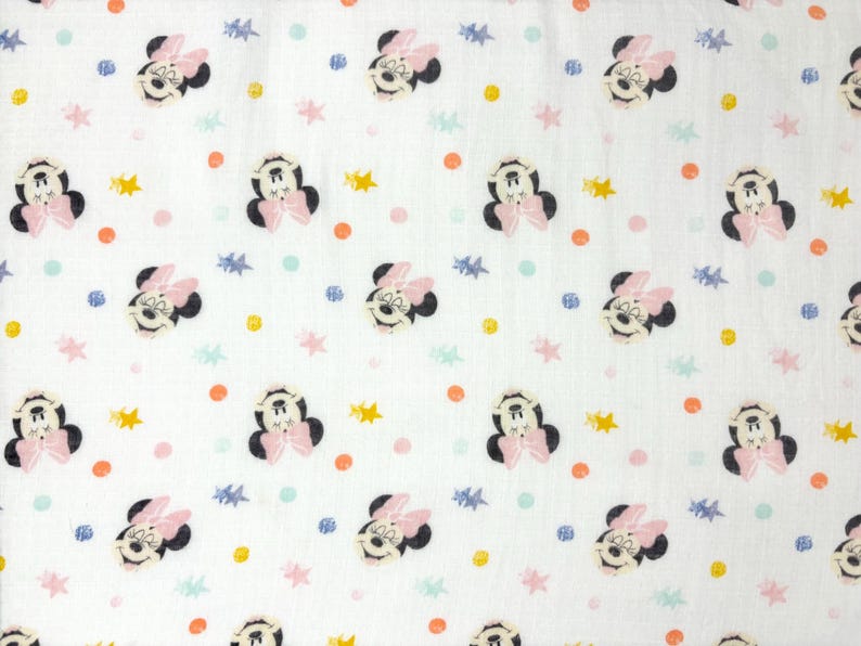 Op de afbeelding: Witte stof met een herhaald patroon van Minnie Mouse-gezichten, roze strikken en kleurrijke stippen en sterren. De Minnie Mouse-gezichten zijn zwart en wit met roze strikken. De stippen zijn oranje, roze en blauw, en de sterren zijn geel en blauw.