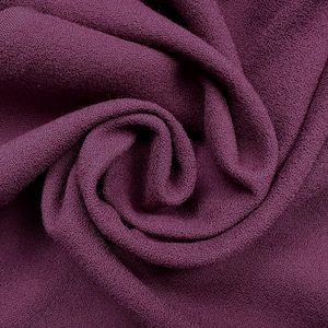 May include: A deep plum purple fleece fabric with a soft, textured surface. The material is draped in a central swirl, showcasing the dense, fuzzy pile and the consistent, rich colour throughout the folds of the textile material.