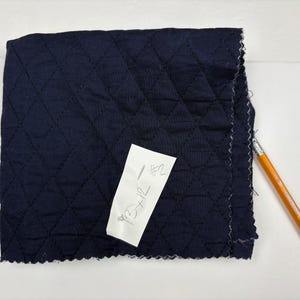 K&ouml;nnte beinhalten: Ein Quadrat aus marineblauem, gestepptem Stoff mit einem Rautenmuster. Ein wei&szlig;es Etikett mit handschriftlichem Text ist angebracht. Ein gelb-oranger Bleistift liegt am Rand des Stoffes.
