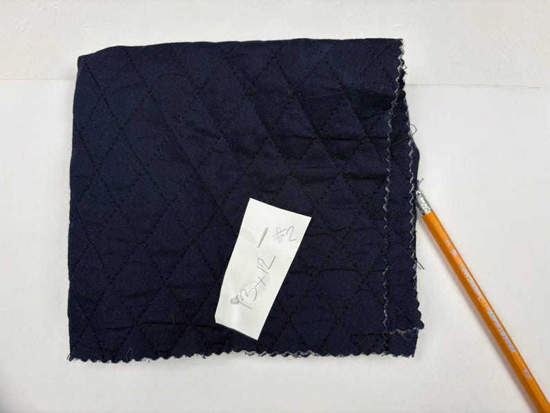 K&ouml;nnte beinhalten: Ein Quadrat aus marineblauem, gestepptem Stoff mit einem Rautenmuster. Ein wei&szlig;es Etikett mit handschriftlichem Text ist angebracht. Ein gelb-oranger Bleistift liegt am Rand des Stoffes.