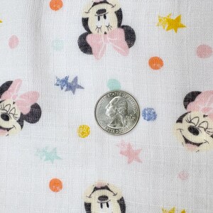 Op de afbeelding: Witte stof met een herhaald patroon van Minnie Mouse gezichten, roze strikken en kleurrijke stippen en sterren. Een Amerikaanse kwart dollar munt is inbegrepen voor de schaal.