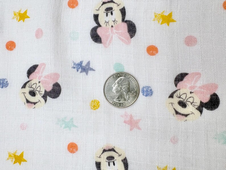 Op de afbeelding: Witte stof met een herhaald patroon van Minnie Mouse gezichten, roze strikken en kleurrijke stippen en sterren. Een Amerikaanse kwart dollar munt is inbegrepen voor de schaal.