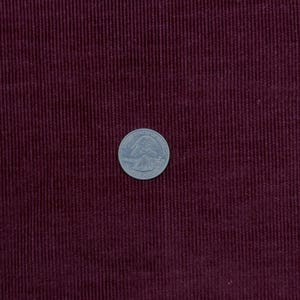 Op de afbeelding: Een diepbordeauxrode corduroy stof met een duidelijke verticale ribbelstructuur. Een zilverkleurige Amerikaanse kwartmunt is in het midden van de stof geplaatst om een duidelijke schaal te bieden voor de breedte van de ribbels.