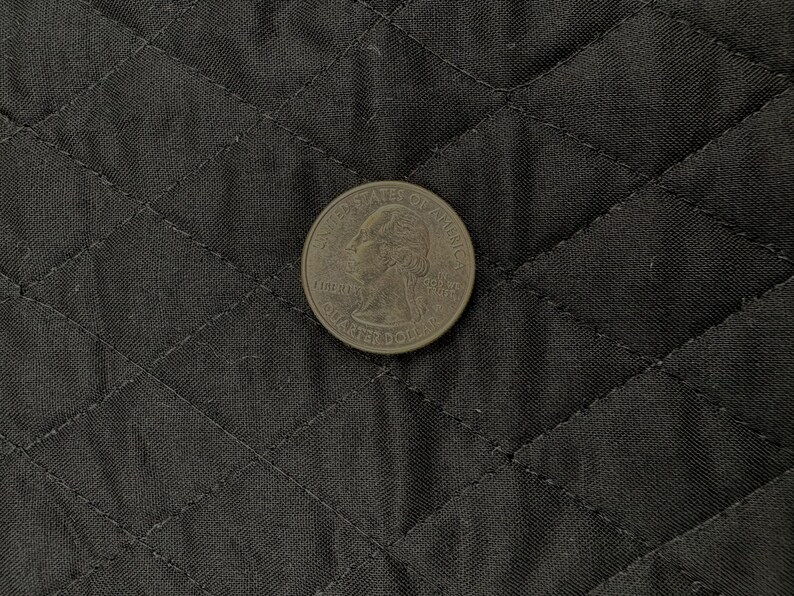 K&ouml;nnte beinhalten: Eine US-amerikanische 25-Cent-M&uuml;nze mit dem Profil von George Washington liegt auf einem schwarzen, gesteppten Stoff. Die Details der M&uuml;nze, einschlie&szlig;lich der Worte "Liberty" und "Quarter Dollar", sind sichtbar. Der Stoff hat ein Rautenmuster.