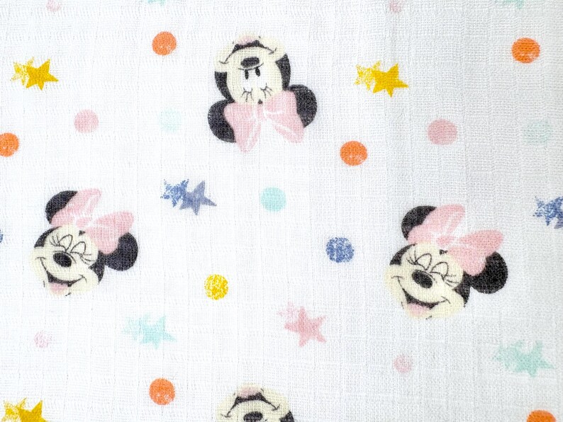 Op de afbeelding: Witte stof met een herhaald patroon van Minnie Mouse-gezichten, roze strikken en kleurrijke stippen en sterren. Het ontwerp heeft gele, oranje, roze, blauwe en groene accenten. De stof heeft een getextureerde uitstraling.