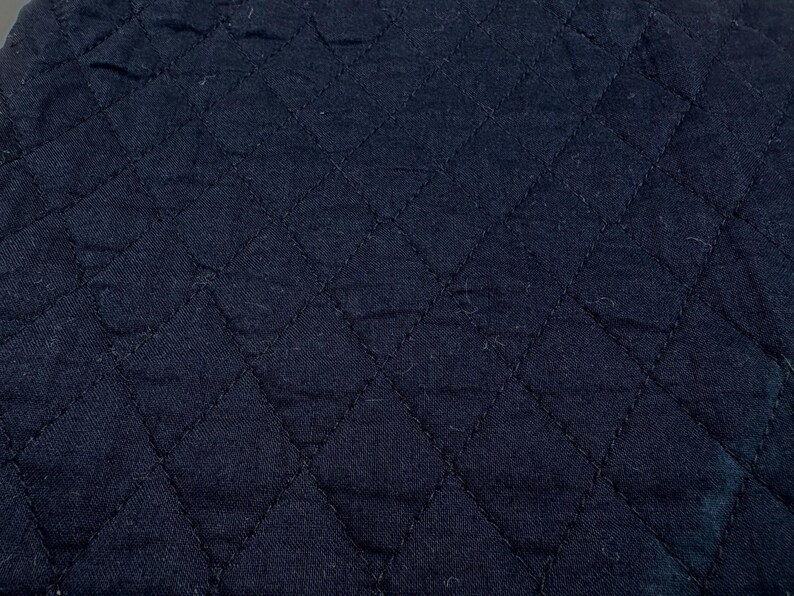 K&ouml;nnte beinhalten: Nahaufnahme eines marineblauen, gesteppten Stoffes. Der Stoff hat ein Rautenmuster, das durch gen&auml;hte Linien erzeugt wird. Die Textur wirkt weich und leicht zerknittert, mit subtilen Farbvariationen.
