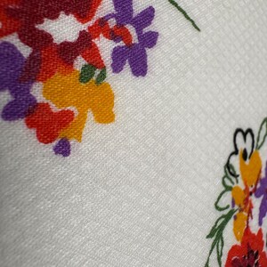May include: Close-up of a white fabric with a floral pattern. The flowers are in shades of red, purple, and yellow, with green stems and leaves. The fabric has a subtle textured pattern.