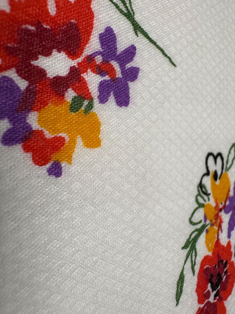 May include: Close-up of a white fabric with a floral pattern. The flowers are in shades of red, purple, and yellow, with green stems and leaves. The fabric has a subtle textured pattern.