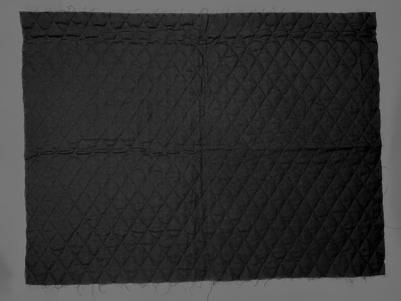 K&ouml;nnte beinhalten: Ein rechteckiges St&uuml;ck schwarzer, gesteppter Stoff. Der Stoff ist mit einem Rautenmuster &uuml;ber die gesamte Oberfl&auml;che gen&auml;ht. Die Kanten des Stoffes sind unfertig, mit sichtbaren losen F&auml;den.