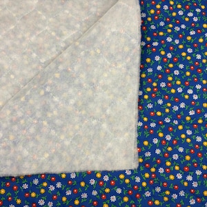 May include: A folded piece of white quilted fabric over a blue floral fabric. The blue fabric has a pattern of small red, yellow, and white flowers. The quilted fabric has a grid pattern.