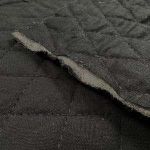 May include: Close-up of black quilted fabric with a diamond pattern. The edge of the fabric is frayed, revealing a lighter gray interior. The fabric has a textured appearance and is likely used for upholstery or crafting.
