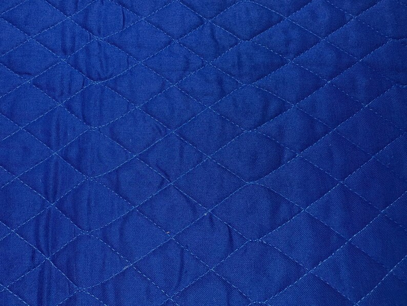 K&ouml;nnte beinhalten: Nahaufnahme eines leuchtend blauen, gesteppten Stoffes. Der Stoff weist ein Rautenmuster auf, das durch gen&auml;hte Linien erzeugt wird. Die Textur wirkt weich und leicht zerknittert, mit subtilen Variationen im Blauton.