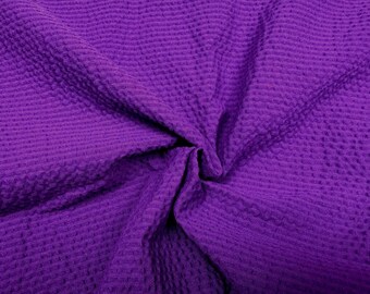 May include: A close-up shot of a vibrant purple fabric with a textured, waffle-like pattern. The fabric is bunched and folded, creating shadows and highlighting the texture. The color is a rich, saturated purple.