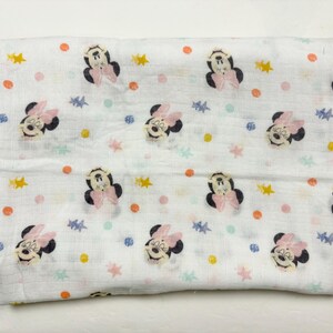 Op de afbeelding: Witte babydeken met een herhaald patroon van Minnie Mouse-gezichten, roze strikken en kleurrijke sterren en stippen. De deken is opgevouwen en ziet er zacht en licht uit, geschikt voor een baby.