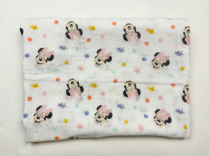 Op de afbeelding: Witte babydeken met een herhaald patroon van Minnie Mouse-gezichten, roze strikken en kleurrijke sterren en stippen. De deken is opgevouwen en ziet er zacht en licht uit, geschikt voor een baby.