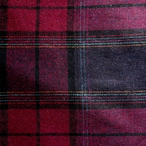 May include: A close-up of a burgundy and black plaid fabric. The pattern features a grid of thick black lines, with burgundy and deep purple squares. Thin lines of teal, gold, and red thread add detail to the design. The fabric has a textured appearance.