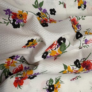 May include: White fabric with a textured surface, adorned with colorful floral patterns. The flowers feature red, yellow, purple, and black petals with green leaves. The fabric is folded, creating shadows and depth.