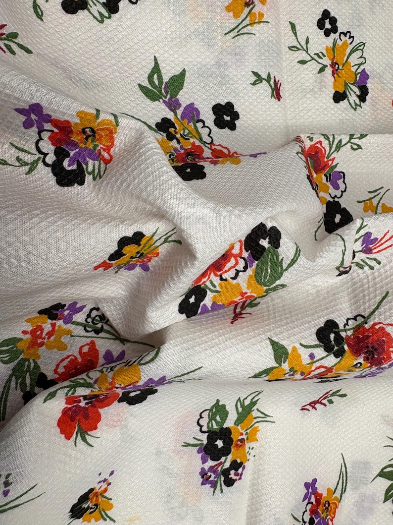 May include: White fabric with a textured surface, adorned with colorful floral patterns. The flowers feature red, yellow, purple, and black petals with green leaves. The fabric is folded, creating shadows and depth.