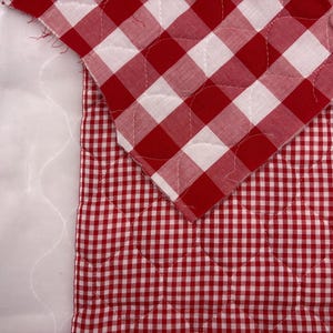 May include: Close-up of a red and white gingham patterned quilt. The quilt features a classic check design, with a quilted pattern visible on the fabric. The edges of the quilt are visible, showing the layers of fabric and batting.
