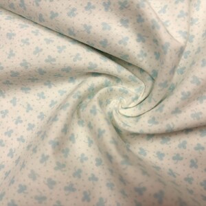May include: Close-up of a white fabric with a repeating pattern of light blue clover-like shapes and small dots. The fabric is gathered in a swirl, showing the texture and design. Ideal for quilting or crafting.