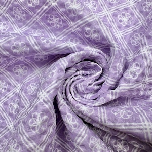 May include: Close-up of a soft, swirled fabric in a light purple hue. The material features a repeating pattern of white floral designs within a grid of white lines, creating a textured, decorative effect. The fabric appears to be lightweight and breathable.
