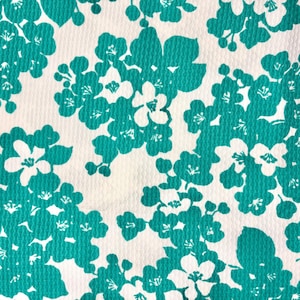 May include: A close-up of a fabric with a repeating floral pattern. The design features teal-colored flowers and clusters of blossoms against a white background. The fabric has a textured appearance.