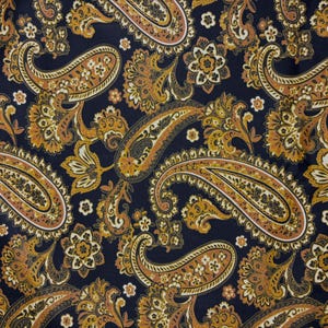 May include: Close-up of a fabric with a repeating paisley pattern. The design features large, ornate paisley shapes in shades of gold, brown, and cream against a dark navy blue background. Small floral accents are scattered throughout the design.