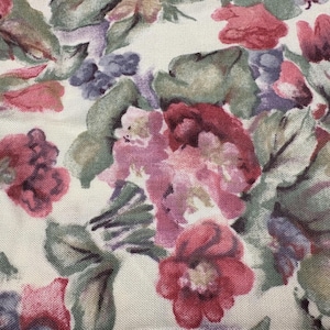 May include: Close-up of a fabric with a floral pattern. The design features a variety of flowers in shades of red, pink, and purple, with green leaves against a cream-colored background. The fabric has a textured appearance.