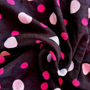 Op de afbeelding: Close-up van een donkere stof met een patroon van roze en witte stippen. De stof is gevouwen, waardoor schaduwen en diepte ontstaan. De stippen variëren in grootte en zijn gelijkmatig verdeeld over het materiaal.
