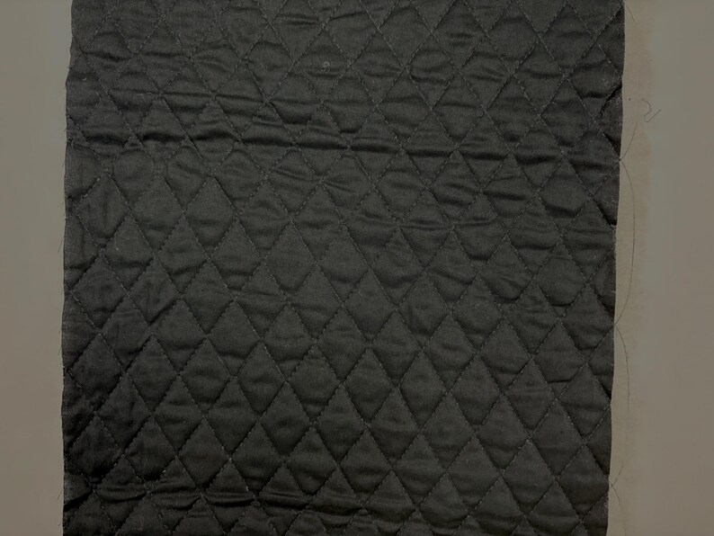 K&ouml;nnte beinhalten: Ein quadratisches St&uuml;ck schwarzes, gestepptes Gewebe mit einem Rautenmuster. Der Stoff hat eine leicht zerknitterte Textur und scheint aus einem weichen Material zu bestehen. Die Steppung erzeugt ein Raster aus Rautenformen auf der Oberfl&auml;che.