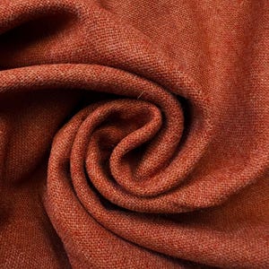 May include: A piece of fabric in a deep, earthy rust-orange color. The material features a visible, dense woven texture with a consistent matte finish. The fabric is arranged in a central spiral fold, creating soft, flowing shadows and highlights.