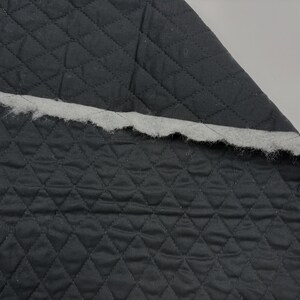 K&ouml;nnte beinhalten: Nahaufnahme eines St&uuml;cks dunkelgrauen, gesteppten Stoffes mit einem Rautenmuster. Der Stoff ist mit einem wei&szlig;en, flauschigen Material an den R&auml;ndern &uuml;berlagert, wodurch ein strukturierter Kontrast entsteht. Der Stoff wird wahrscheinlich f&uuml;r Polsterarbeiten oder Bastelarbeiten verwendet.