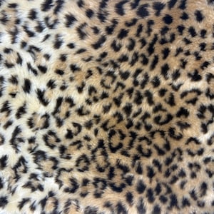 May include: A soft, textured fabric featuring a repeating leopard print pattern. The design consists of irregular black spots with brown centers arranged against a light tan and cream-colored background, creating a dense, fur-like appearance.
