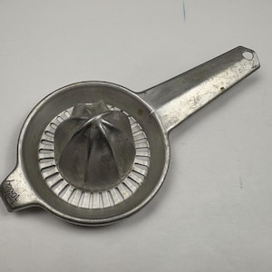 May include: A vintage, silver-toned citrus juicer with a long handle and a reamer. The juicer has a circular bowl with a ridged interior and a spout. The handle has a hole for hanging. The word "Toby" is stamped on the side.