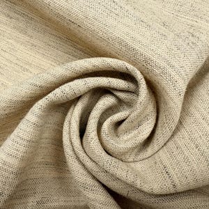 May include: A beige fabric with a visible woven texture and subtle dark flecks throughout. The material is gathered into a central spiral fold, highlighting the depth of the weave and the natural, light tan color of the textile fibers.