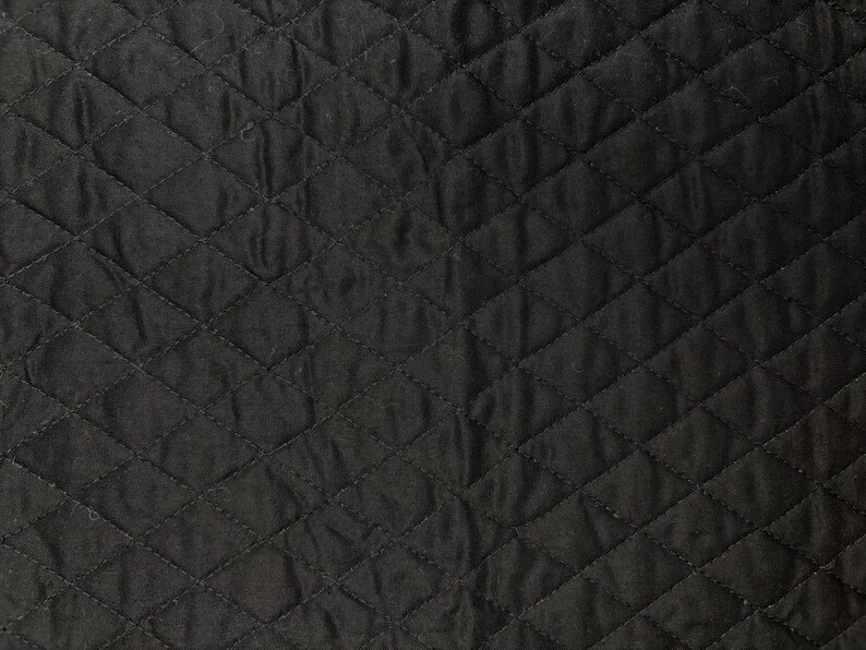 K&ouml;nnte beinhalten: Nahaufnahme eines schwarzen, gesteppten Stoffes mit einem Rautenmuster. Der Stoff hat eine leicht zerknitterte Textur, und die N&auml;hte erzeugen ein gitterartiges Design. Die dunkle Farbe und Textur deuten auf ein strapazierf&auml;higes Material hin.