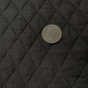 K&ouml;nnte beinhalten: Eine Vierteldollarm&uuml;nze liegt auf einem schwarzen, gesteppten Stoff. Der Stoff hat ein Rautenmuster, das durch gen&auml;hte Linien erzeugt wird. Die M&uuml;nze ist silberfarben und zeigt das Profil einer Person.