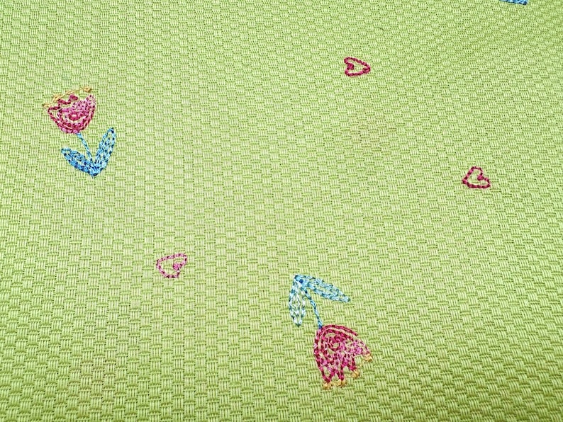 May include: A light green fabric with a textured basketweave pattern featuring small embroidered motifs. The design includes pink and blue tulips with green stems and scattered pink heart outlines stitched across the surface of the material.